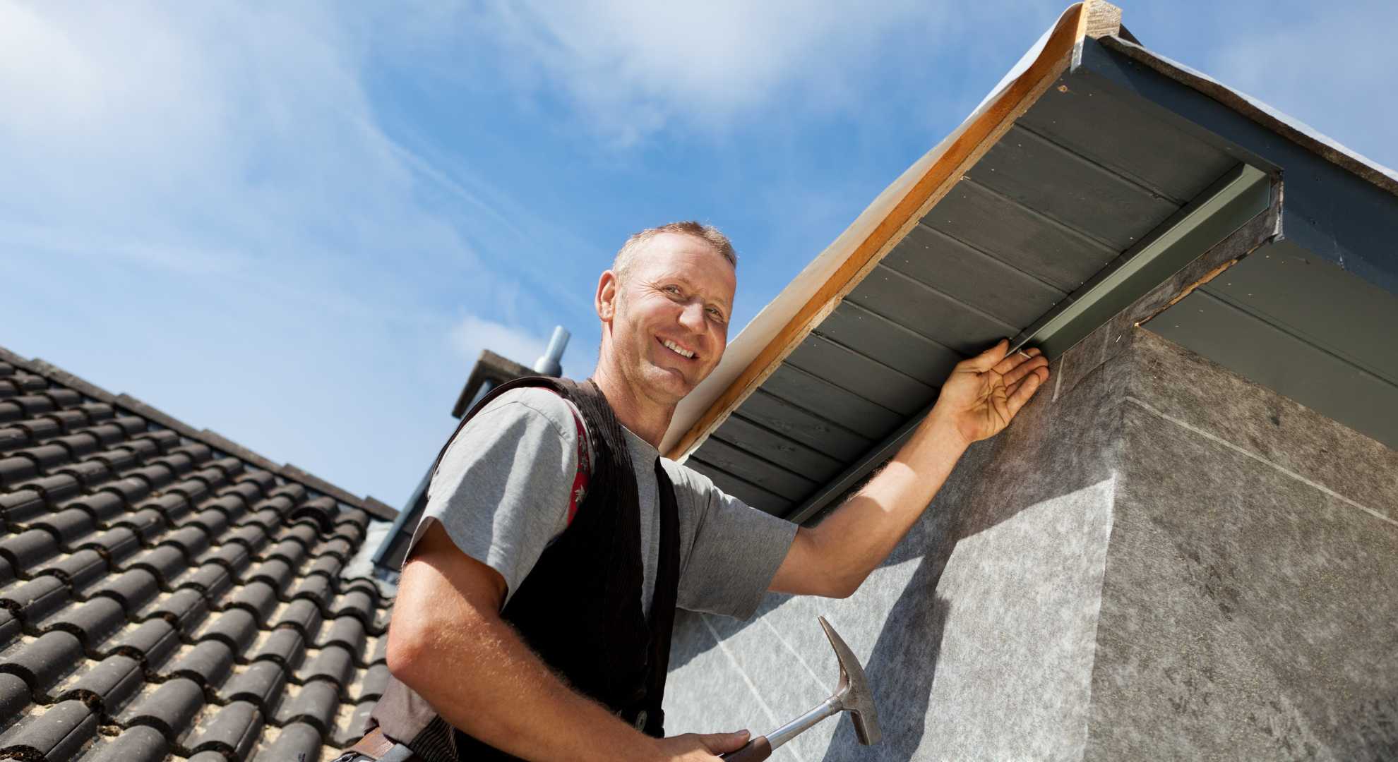 Dachdecker1.jpg Dachdecker bei der Arbeit an der Dachkante eines Hauses, lächelnd mit Hammer in der Hand unter blauem Himmel.