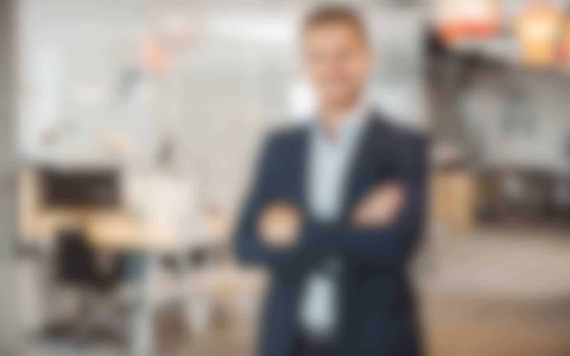 CEO-Gross-neu.jpg Mann im Anzug mit verschränkten Armen in einem modernen Büro, unscharf im Hintergrund dargestellt.