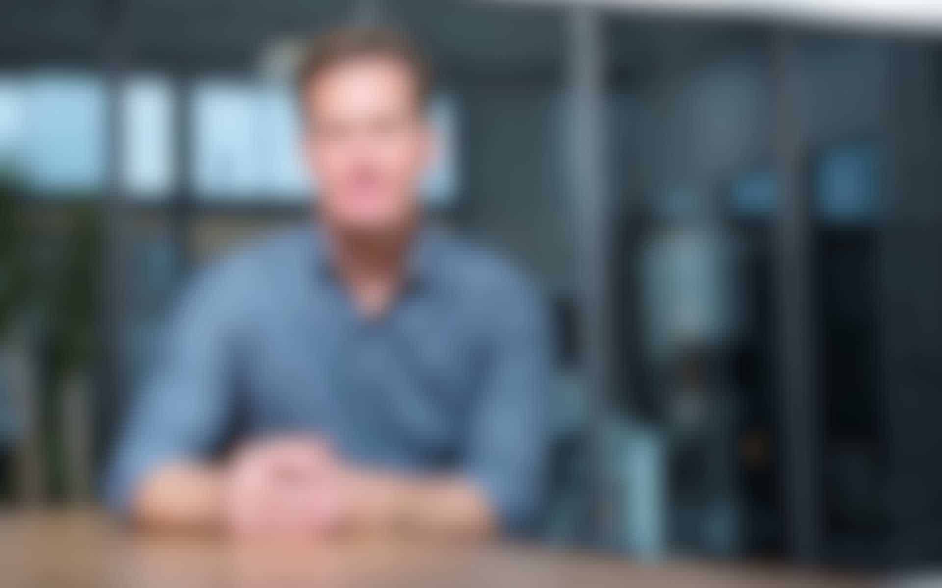 IT-Gross.jpg Unscharfes Porträt eines Mannes mit blauem Hemd, der an einem Tisch in einem modernen Büro sitzt.