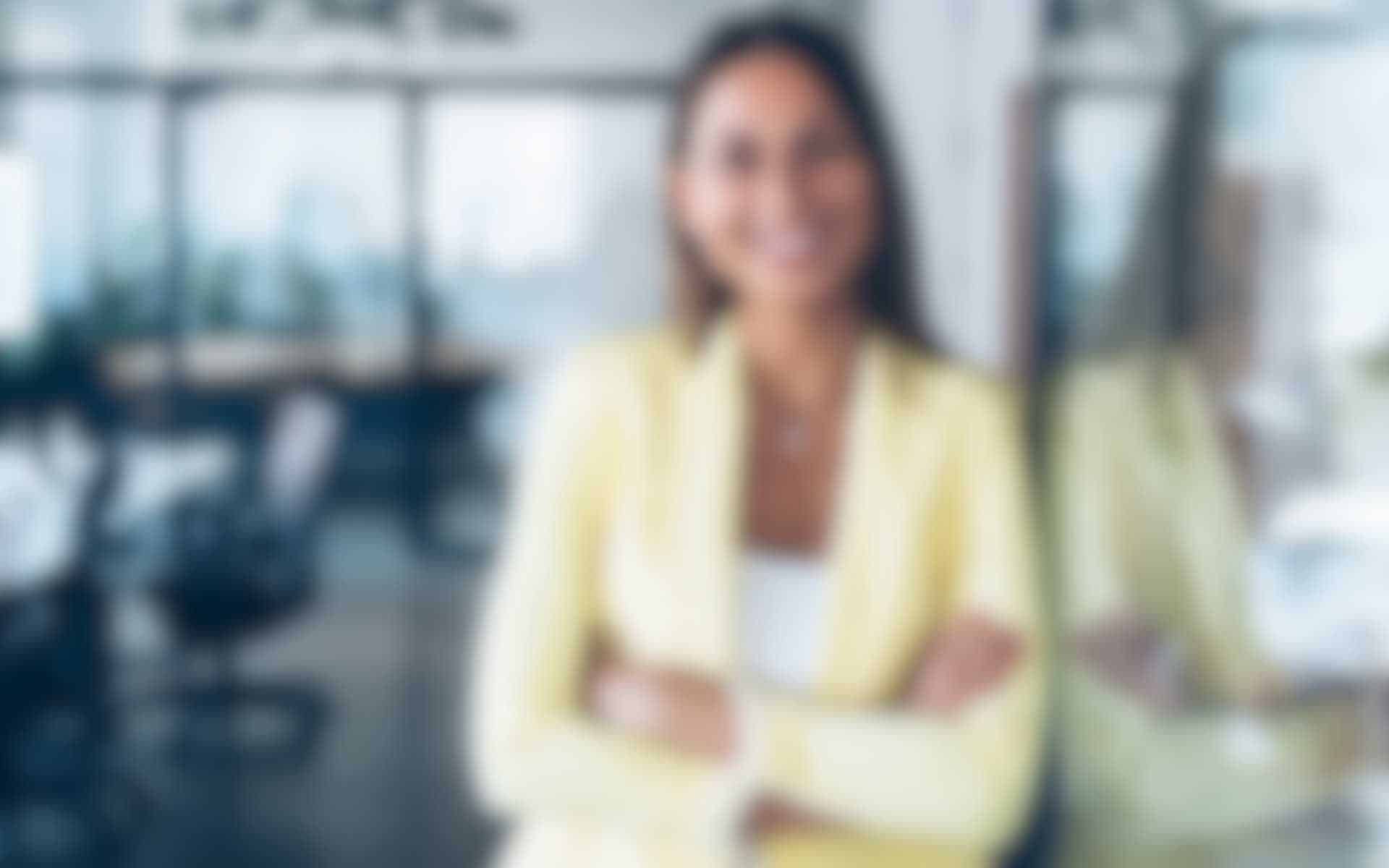 Marketing-Gross.jpg Junge Geschäftsfrau mit langem braunem Haar und gelbem Blazer steht mit verschränkten Armen lächelnd vor einer Glaswand in einem modernen Büro.