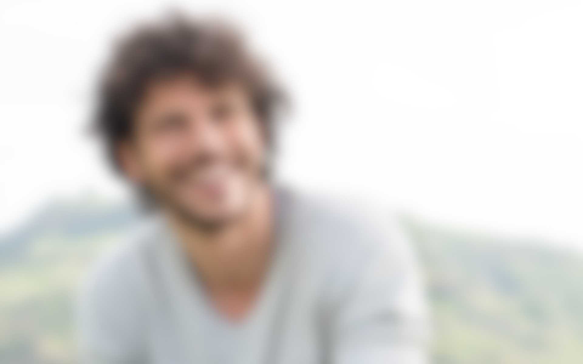 Strategie-Experte-Gross.jpg Unscharfes Bild eines lächelnden Mannes mit lockigem Haar im Freien vor einer hügeligen Landschaft.