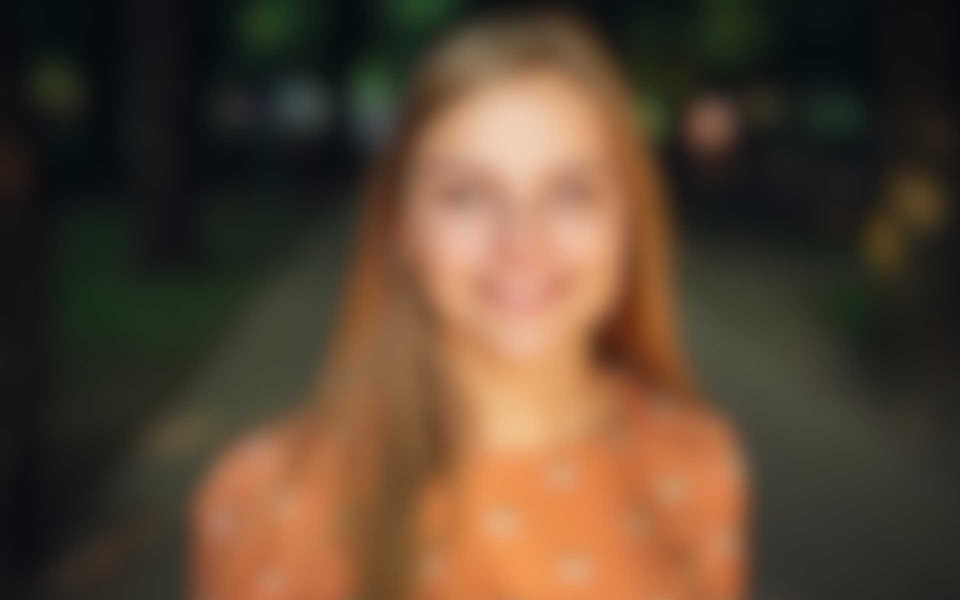 Support-Klein.jpg Unscharfes Porträt einer jungen Frau mit langen Haaren in einem orangefarbenen Kleid, lächelnd im Freien auf einem Weg im Park.