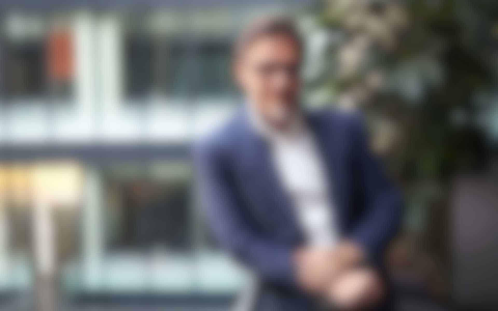 Unternehmensberater-Gross.jpg Unscharfes Porträt eines lächelnden Geschäftsmannes im blauen Sakko vor einem modernen Bürogebäude.