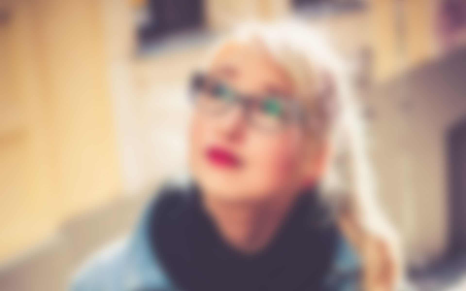 team_4.jpg Porträt einer Frau mit Brille und blondem Zopf, die nachdenklich nach oben blickt. Der Hintergrund zeigt eine städtische Umgebung.