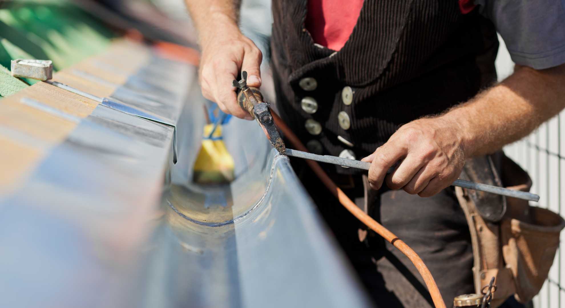 Leckage.jpg Handwerker bei der Reparatur einer Dachrinne mit einem Lötkolben – Detailaufnahme der präzisen Leckabdichtung.