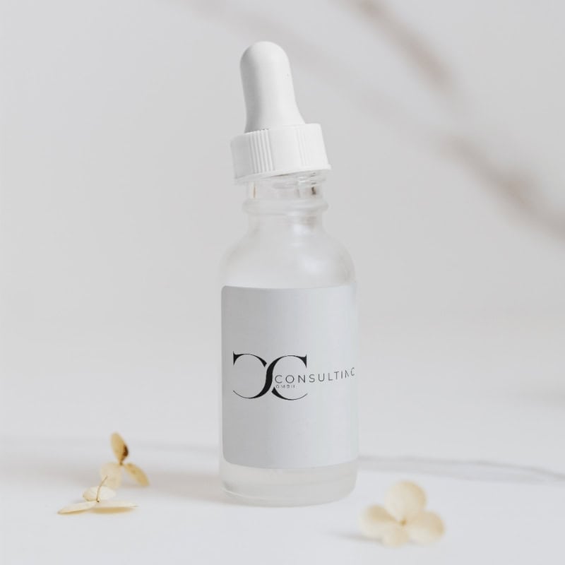 Transparente Pipettenflasche mit weißem Etikett und dem Logo „CC Consulting GmbH“ auf hellgrauem Hintergrund. Die Flasche steht auf einer weißen Oberfläche, dekoriert mit getrockneten Blüten.