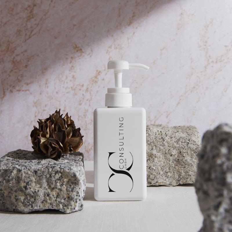 Eleganter weißer Pumpspender mit schwarzem Logo „CC Consulting GmbH“, stehend auf hellem Untergrund zwischen Natursteinen und dekorativer Trockenblume. Der Hintergrund ist marmoriert in zarten Rosatönen.