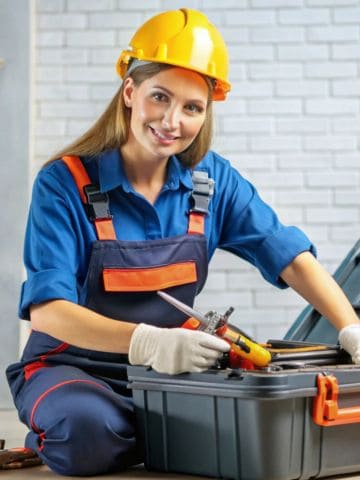 Eine Handwerkerin in blauer Arbeitskleidung mit orangefarbenen Akzenten und gelbem Schutzhelm sitzt vor einem offenen Werkzeugkoffer. Sie trägt Arbeitshandschuhe und hält lächelnd ein Werkzeug in der Hand. Im Hintergrund ist eine helle Ziegelwand zu sehen.