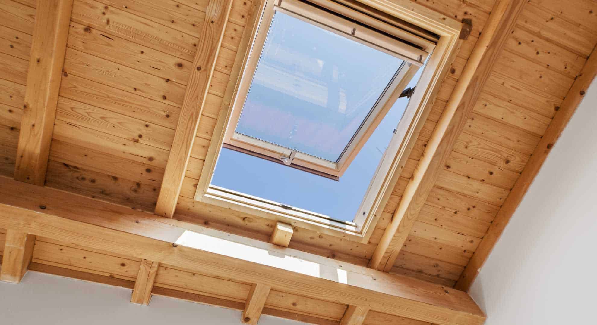 Blick von innen auf ein geöffnetes Dachfenster in einer Holzdecke mit sichtbaren Balken. Durch das Fenster ist ein klarer blauer Himmel zu sehen, und Sonnenlicht fällt in den Raum.