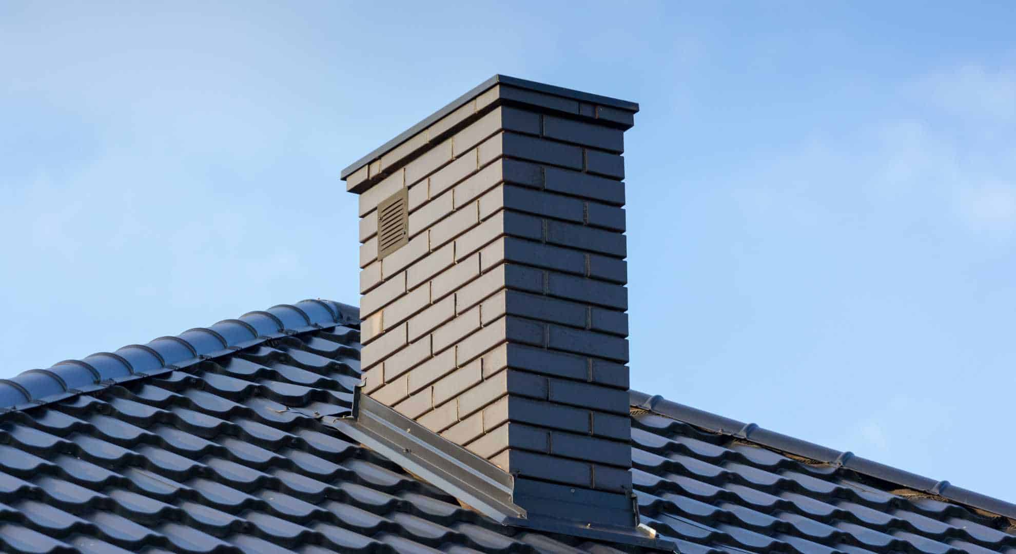 Ein moderner gemauerter Schornstein aus dunklen Klinkersteinen ragt aus einem Dach mit glänzenden, schwarzen Dachziegeln. Der Himmel ist klar und hellblau.