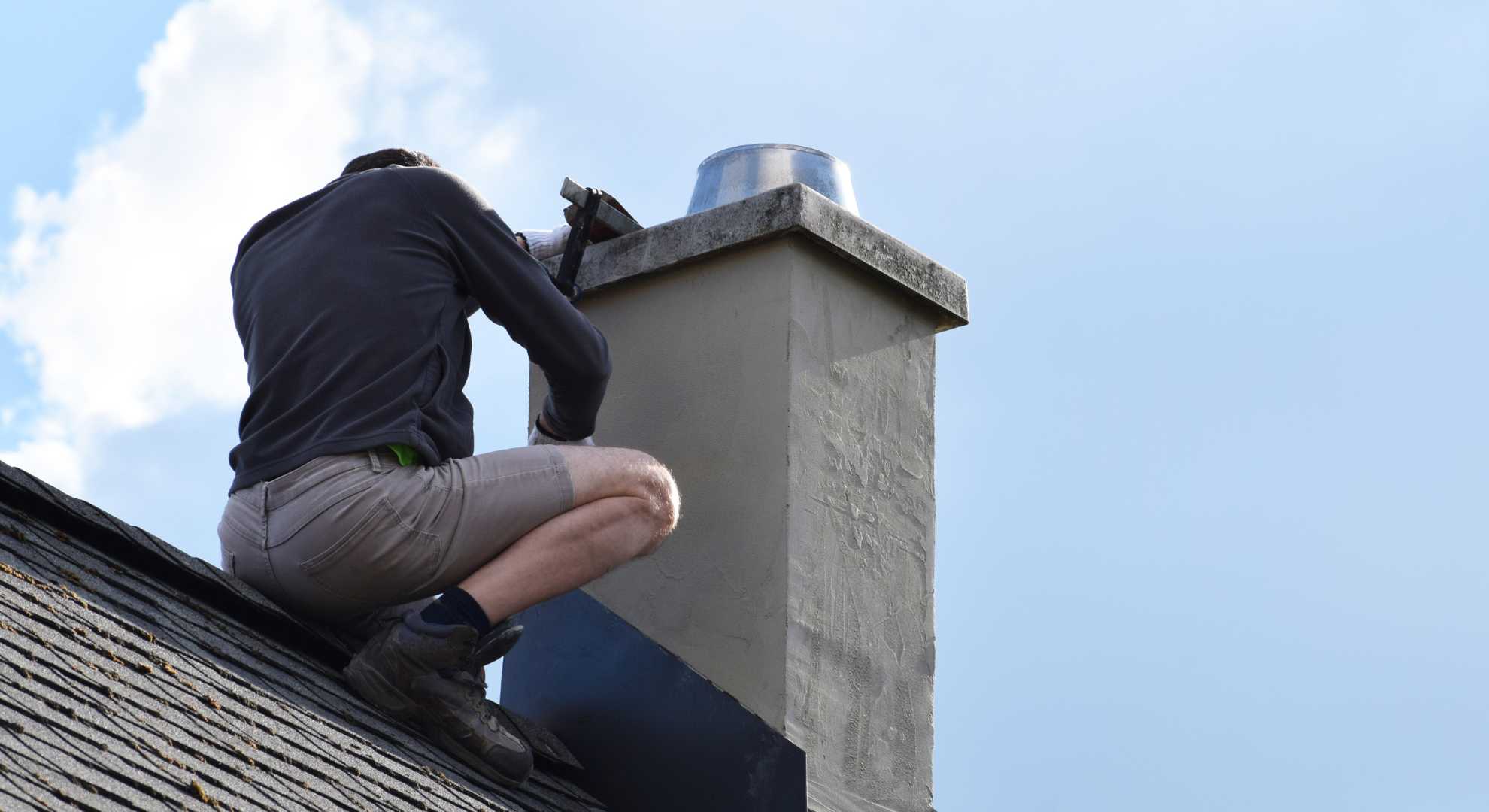 Ein Handwerker in Arbeitskleidung kniet auf einem geneigten Dach und führt Reparaturarbeiten an einem verputzten Schornstein durch. Er verwendet eine Kelle, während der Himmel im Hintergrund leicht bewölkt ist.
