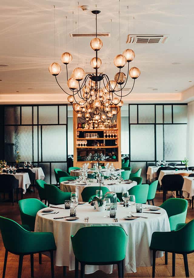Elegantes Restaurant mit runden, weiß gedeckten Tischen, grünen Stühlen und modernen Kronleuchtern, im Hintergrund ein Weinregal und stilvolle Raumteiler.
