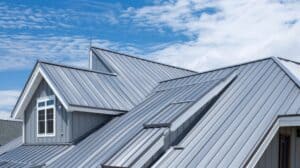 Detailansicht eines modernen Hauses mit grauem Metalldach, Giebelfenstern und mehreren Dachschrägen unter blauem Himmel mit lockerer Bewölkung.