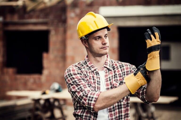 Bauarbeiter mit gelbem Schutzhelm und kariertem Hemd zieht sich auf einer Baustelle robuste Arbeitshandschuhe an. Im Hintergrund ist ein Rohbau mit offenen Fenstern und Baugeräten zu sehen.