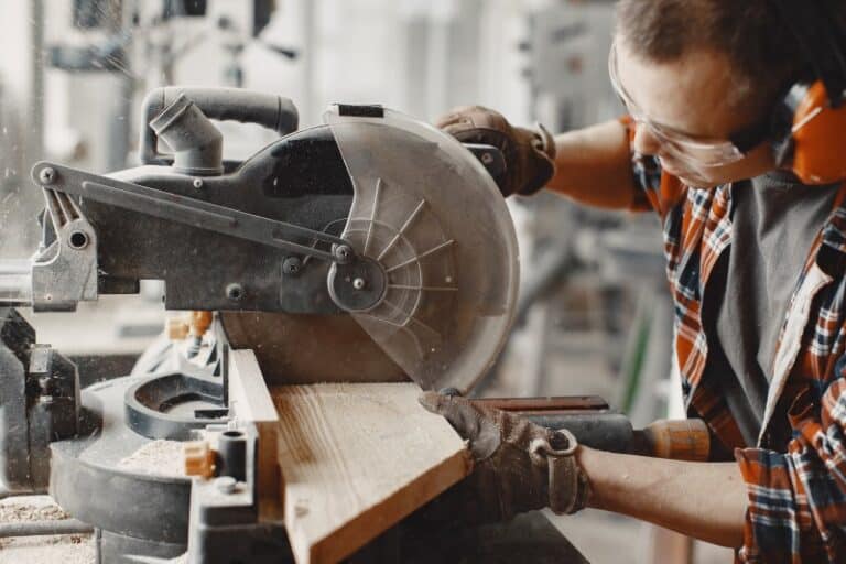 Handwerker mit Schutzbrille, Gehörschutz und Handschuhen arbeitet konzentriert mit einer Kreissäge und schneidet ein Holzbrett in einer Werkstatt.