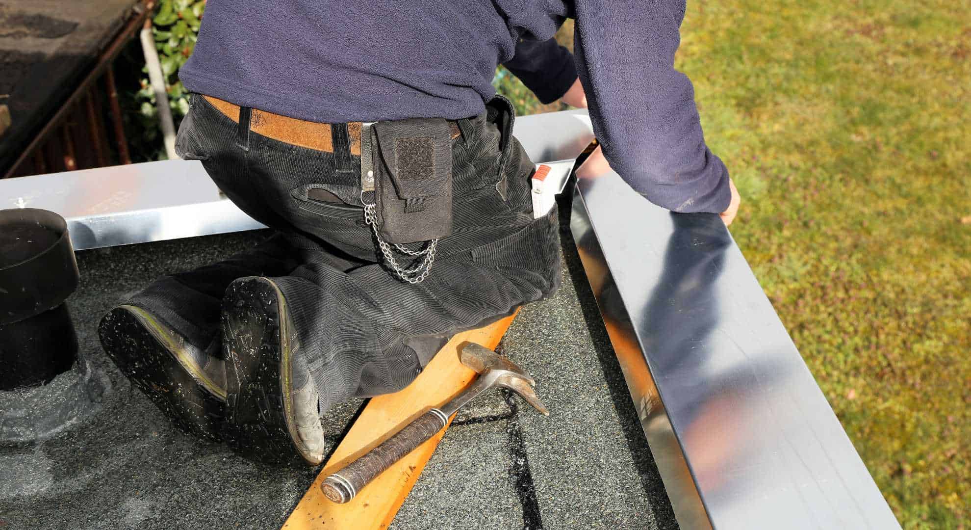 Ein Handwerker kniet auf einem Flachdach und montiert eine Metallabdeckung am Dachrand. Neben ihm liegen ein Hammer und ein Holzkeil. Er trägt robuste Arbeitskleidung und Werkzeugtasche, im Hintergrund ist eine Rasenfläche zu sehen.