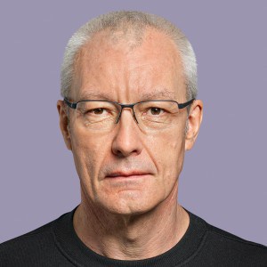 Eingeschränkte Nahaufnahme eines ernst blickenden älteren Mannes mit Brille in neutralem Hintergrund.