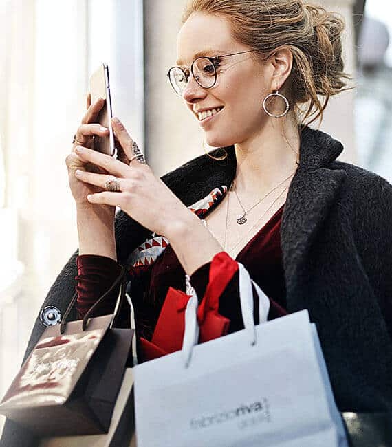 Lächelnde Frau mit Brille und Einkaufstüten schaut auf ihr Smartphone, stilvoll gekleidet beim Shopping.