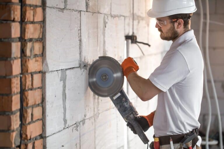 Handwerker mit Schutzhelm, Schutzbrille und Handschuhen schneidet mit einer Trennscheibe präzise in eine Mauer aus Porenbetonsteinen. Er trägt einen Werkzeuggürtel und arbeitet auf einer Baustelle im Rohbau.