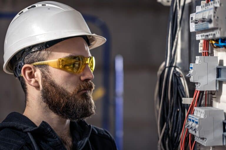 Elektriker mit weißem Schutzhelm und gelber Schutzbrille arbeitet an einem Schaltschrank mit zahlreichen Kabeln und Sicherungseinheiten. Der Fokus liegt auf konzentriertem Arbeiten und Sicherheit.
