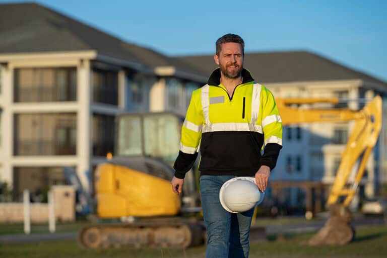 Baustelle eines Hochhauses mit eingerüsteten Gebäuden und mehreren Turmdrehkränen vor einem dramatischen Abendhimmel. Die Konstruktion ist von Stahlgerüsten und grünem Netzschutz umgeben.
