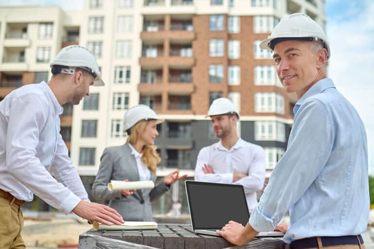 Ein Team von Architekten oder Bauingenieuren mit Schutzhelmen diskutiert auf einer Baustelle vor einem modernen Wohngebäude. Im Vordergrund steht ein Mann mit Laptop, der lächelnd in die Kamera blickt.