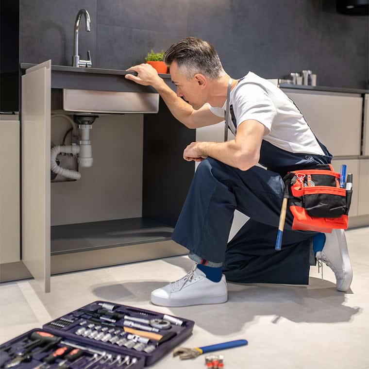 Ein Installateur kniet vor einem offenen Küchenschrank und inspiziert die Abflussrohre unter der Spüle. Er trägt blaue Arbeitskleidung, ein weißes T-Shirt und hat eine Werkzeugtasche am Gürtel. Vor ihm liegt ein geöffneter Werkzeugkoffer mit diversen Werkzeugen auf dem Boden.