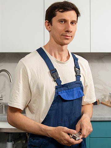 Ein Handwerker in blauer Latzhose und weißem T-Shirt steht in einer modernen Küche vor einer Spüle. Er hält ein kleines Werkzeug in den Händen und blickt ruhig in die Kamera.