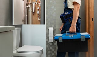 Ein Klempner in Arbeitskleidung betritt ein modernes Badezimmer mit Werkzeugkoffer in der Hand. Im Vordergrund ist eine Toilette zu sehen, im Hintergrund ein geöffneter Technikschrank mit sichtbaren Rohrleitungen und Ventilen.