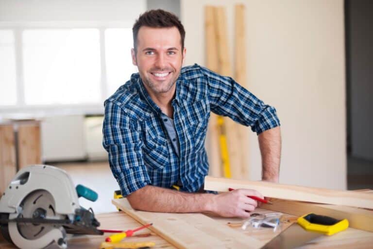 Lächelnder Handwerker in kariertem Hemd arbeitet an einem Holztisch in einer Werkstatt. Um ihn herum liegen Werkzeuge wie eine Kreissäge, Maßband, Bleistift und Schrauben.