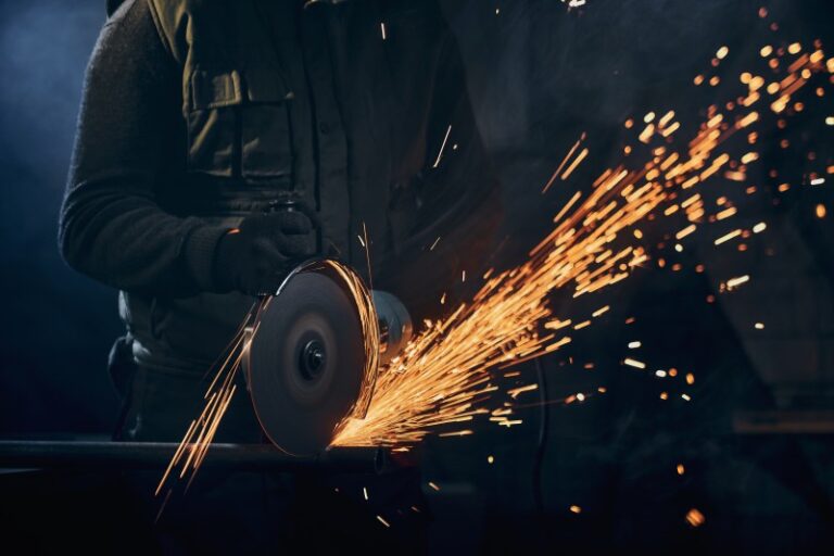 Arbeiter mit Schutzkleidung und Handschuhen schleift ein Metallstück mit einer Trennscheibe. Funken sprühen hell in einem dunklen Arbeitsumfeld – Symbol für Metallverarbeitung oder Industriearbeit.