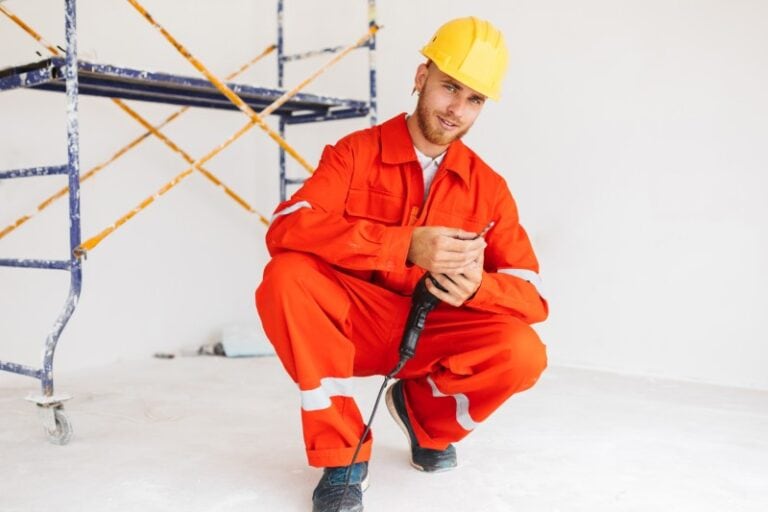 Junger Bauarbeiter in orangefarbener Arbeitskleidung und gelbem Schutzhelm hockt auf dem Boden, hält einen Bohrer in den Händen und lächelt in die Kamera. Im Hintergrund ist ein Baugerüst zu sehen.