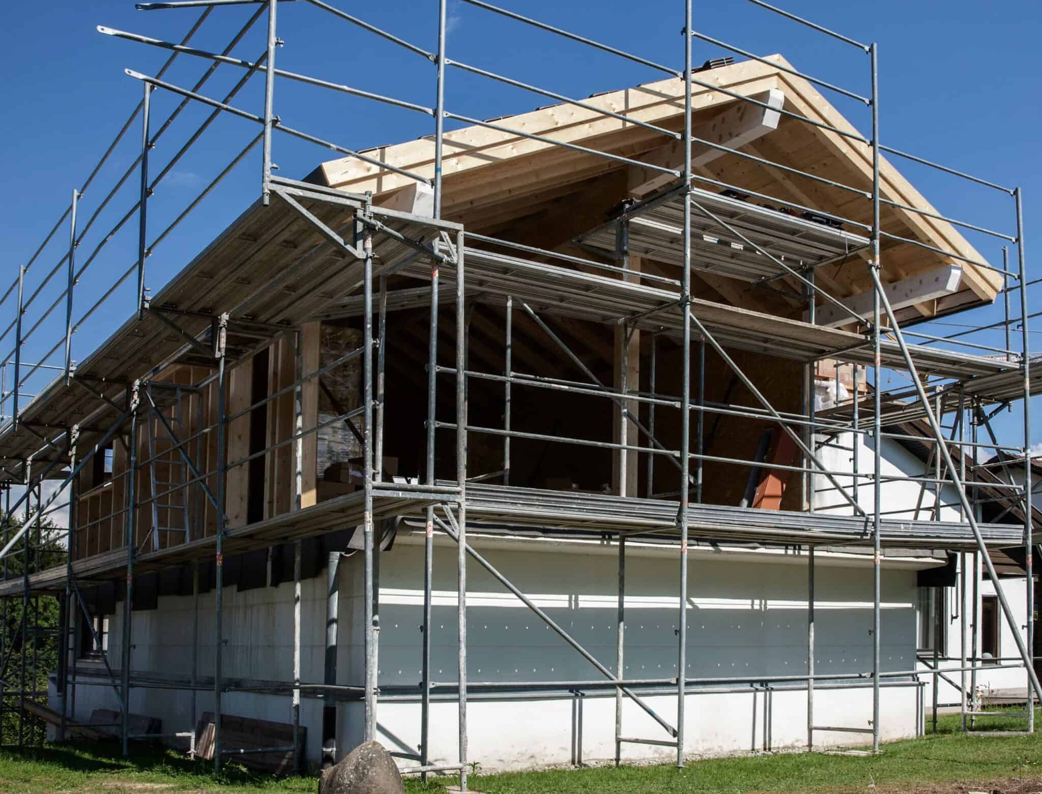 Ein teilweise fertiggestelltes Haus mit Holzrahmenbauweise, eingerüstet mit einem umfangreichen Gerüstsystem, unter blauem Himmel.